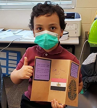 Young student wearing a mask holding up his project